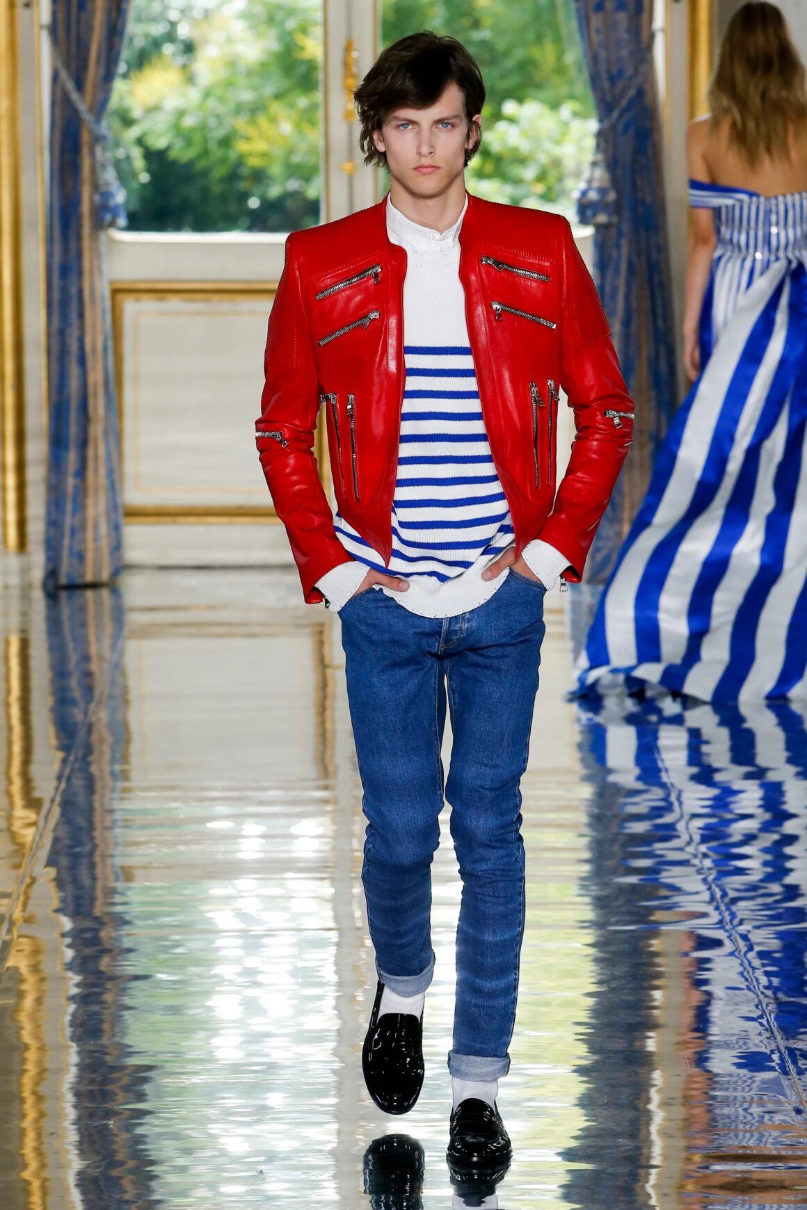 Male model wearing a blue and white stripe shirt, red jacket with zippers, denim pants, and black shoes