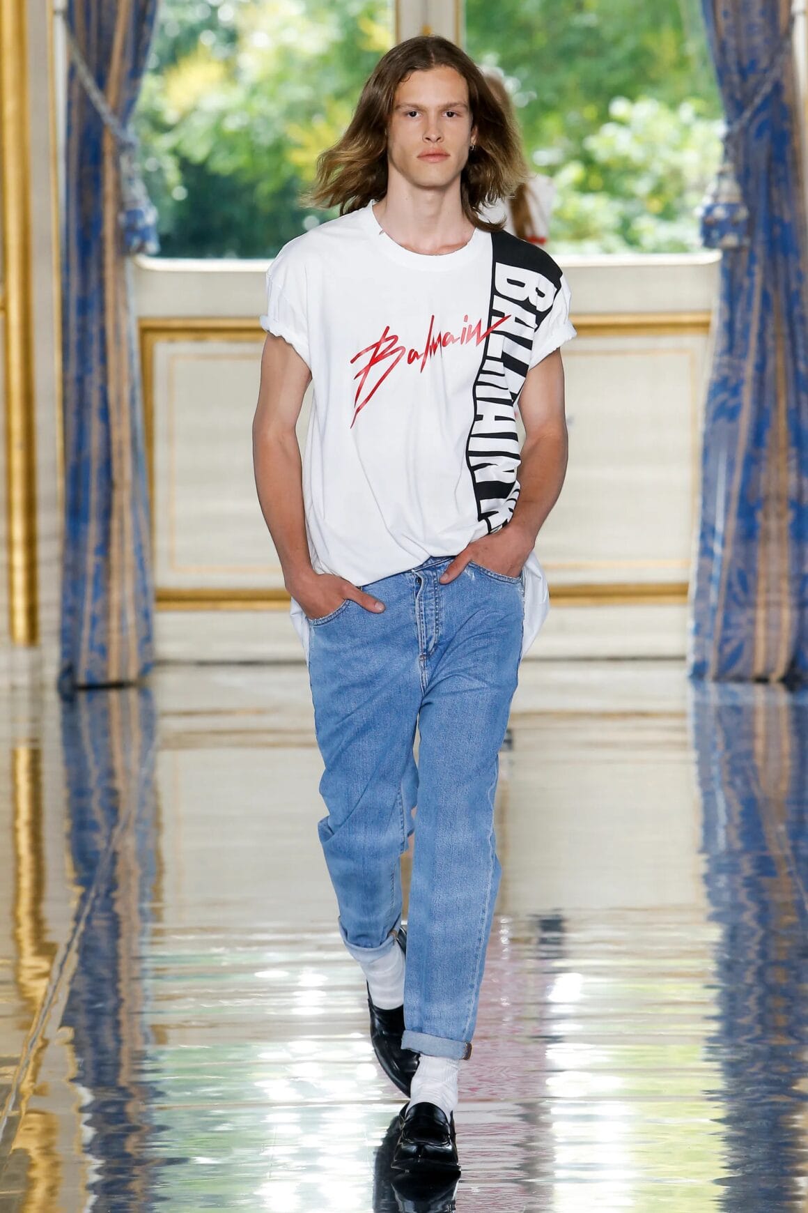 Male model wearing a white black and red tee with Balmain graphic, jeans, black shoes, and white socks