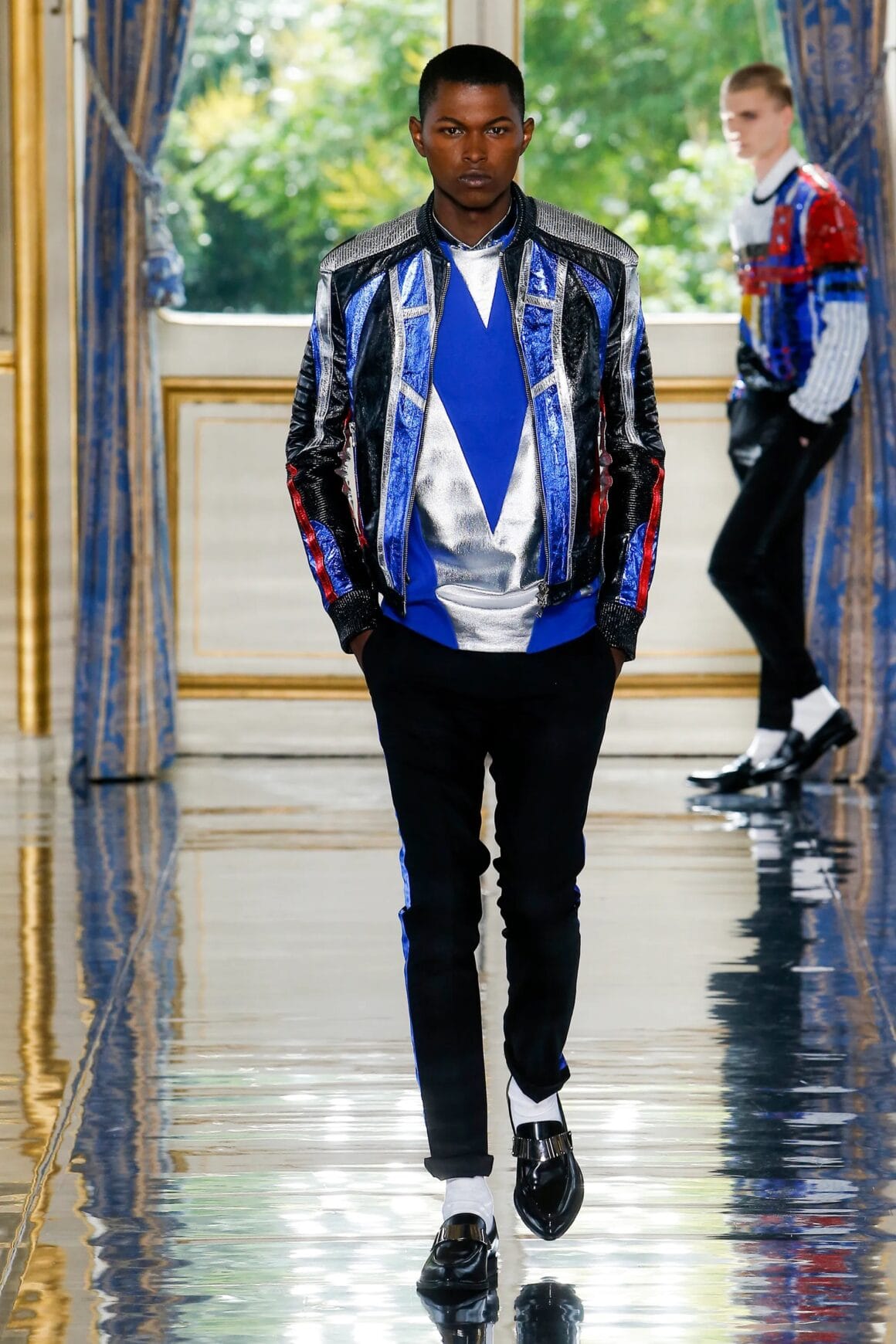 Male model wearing a blue and metallic shirt with a black, metallic, and blue jacket, black pants, and black shoes