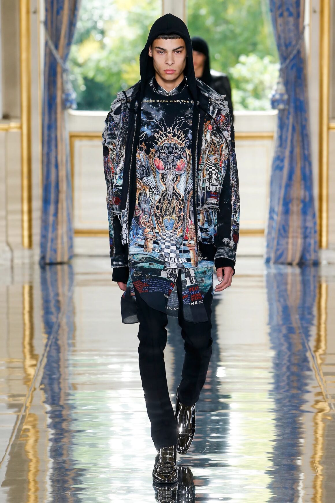 Male model wearing a hooded graphic jacket over a colorful graphic shirt with black pants and black metallic shoes