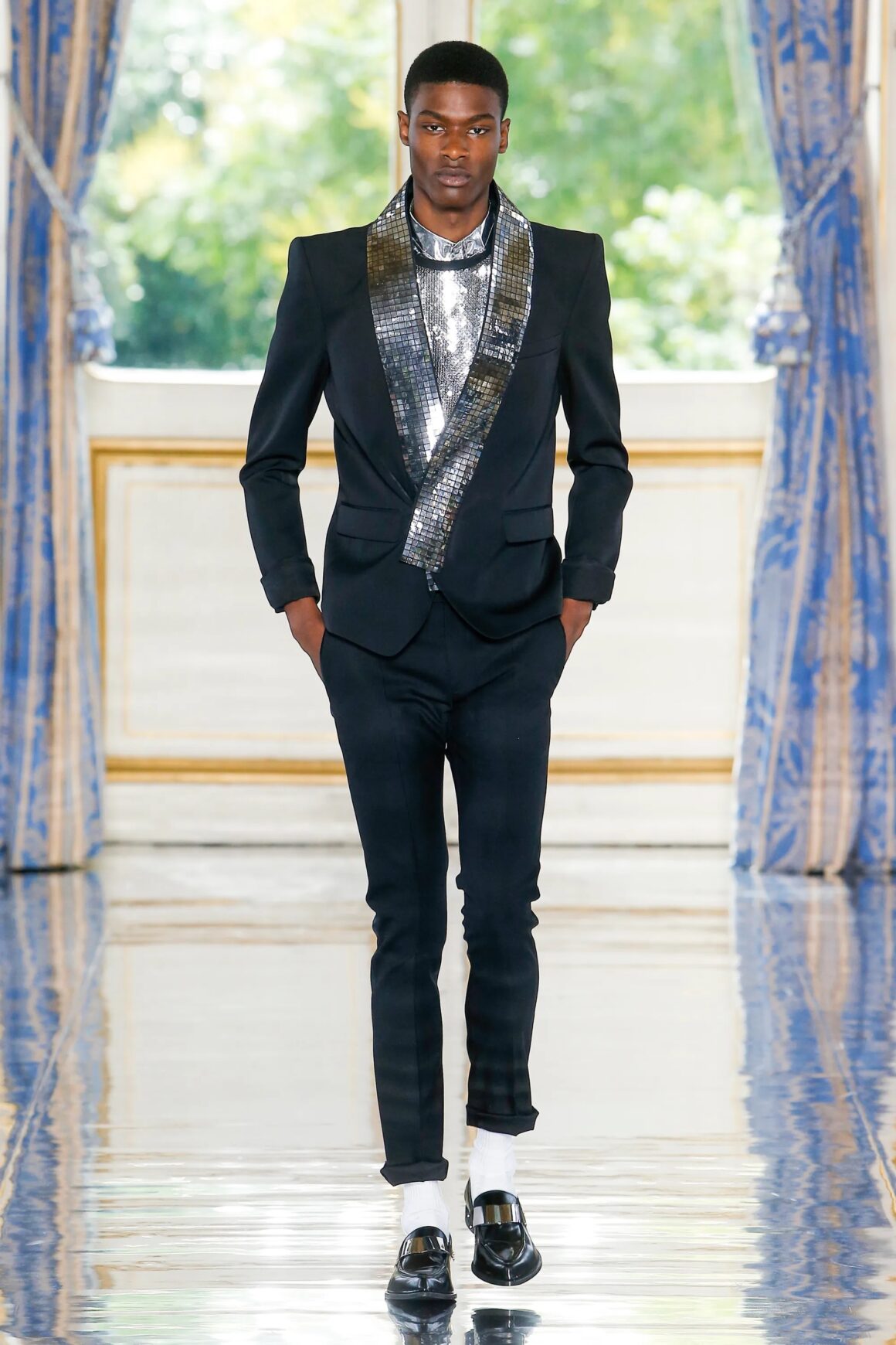 Male model wearing a black and metallic tuxedo jacket with black pants, black shoes, and white socks