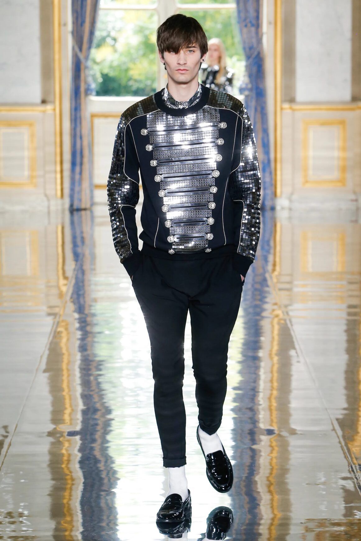 Male model wears black and metallic military style jacket with black pants and black penny loafers with white socks