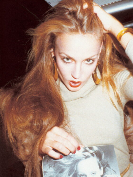 Model Jerry Hall wearing large hoop earrings and matching bracelet, a camel colored sweater, holding a picture of herself