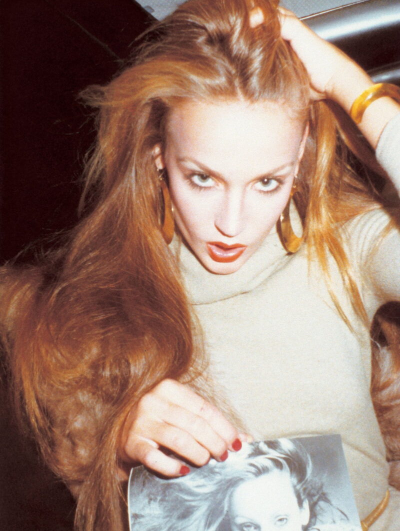 Model Jerry Hall wearing large hoop earrings and matching bracelet, a camel colored sweater, holding a picture of herself